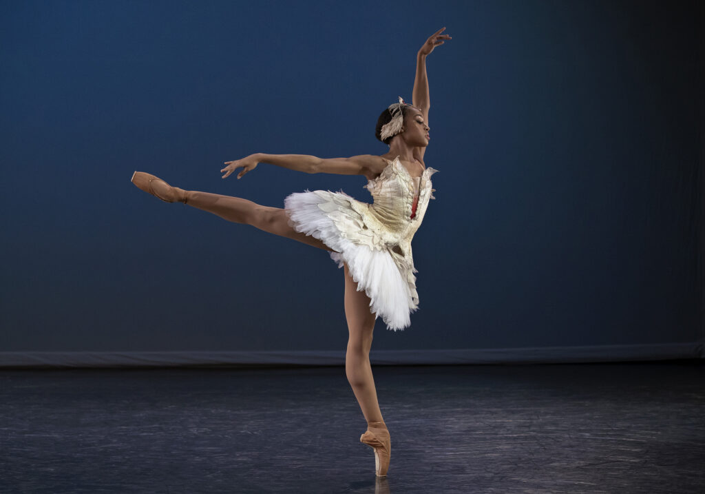 Aleisha Walker poses in first arabesque facing stage left, her eyes downcast and her left arm raised high along the left side of her face. She wears a white tutu with feather embellishments, a feathered headpiece, brown tights and brown pointe shoes.