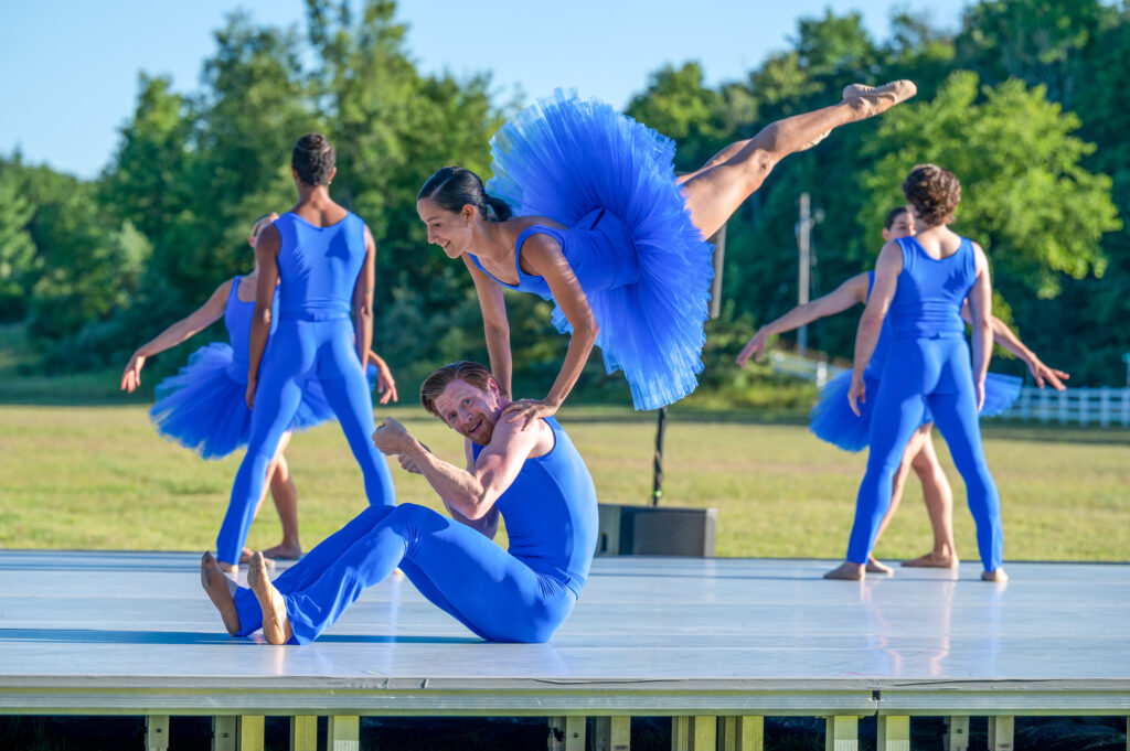 Three couples wearing bright blue costumes perform on an outdoor stage. Two couples stand at the back of the stage, facing each other as they pose. In the foreground, and woman jumps up, her body almost vertical with her legs shotting out behind her, as she leans her hands on her male partner's shoulders as he sits on the ground, looking surprised. The women wear leotards and tutus, while the men wear tank tops and stretchy pants.