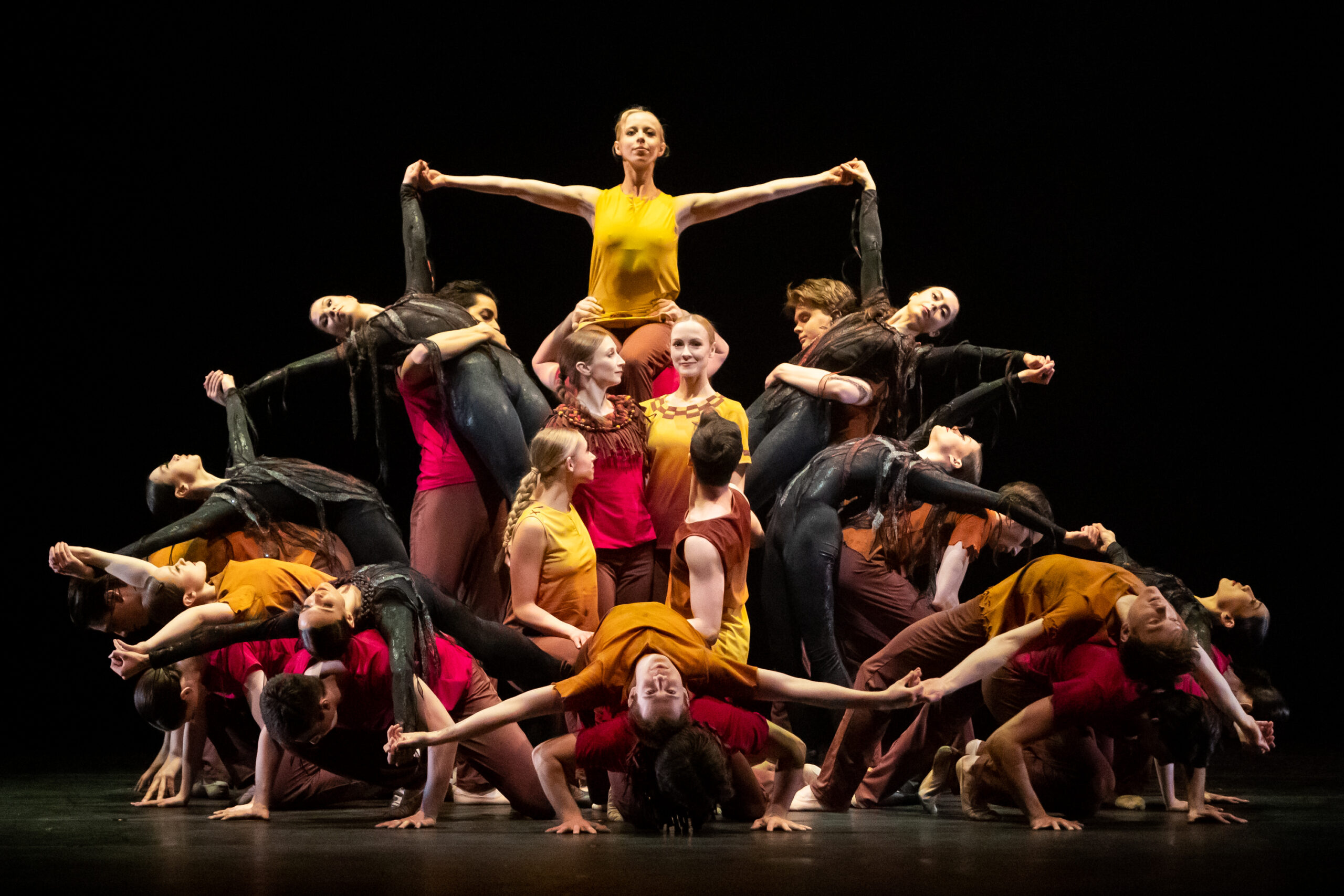 A large group of dancers wearing black, brown, red, orange, and yellow costumes create a symmetrical multi-layer formation by lifting each other up, leaning back, laying on top of each other, and standing in the middle.