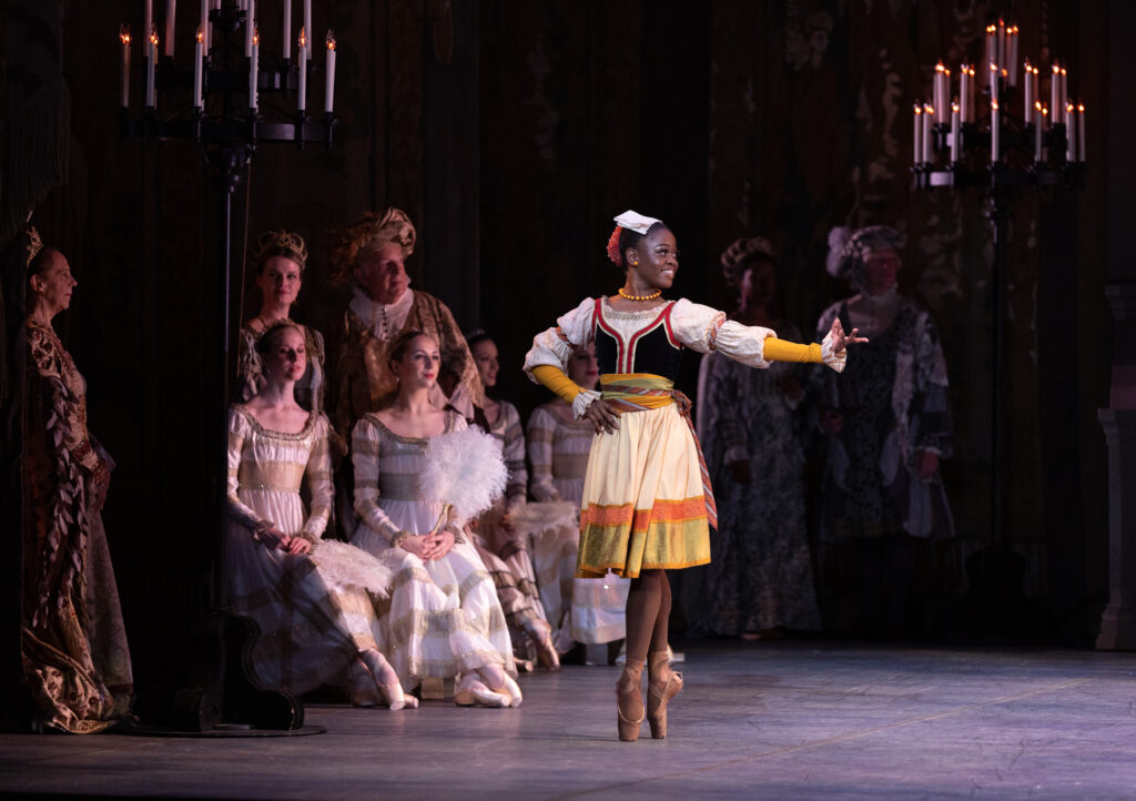 Michaela Mabinty DePrince poses in sous-sus onstage with her right hand on her hip and her left arm extended out to the side. She looks over her left should and smiles. She wears a multi-colored, knee-length dress, brown pointe shoes and a red and white headpiece. Other dancers in aristocratic, medieval costumes sit on stools behind her and watch.