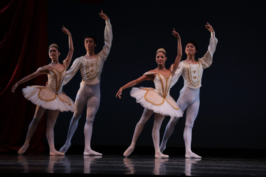 Two male and femal couples stand in tendu derriere effacé with their left arms up in fifth and their right arms in demi-second. The women wear white tutus with pink appliqués and tiars, with pink tights na dpointe shoes. The men wear white tunics with gold trim with white tights and slippers.