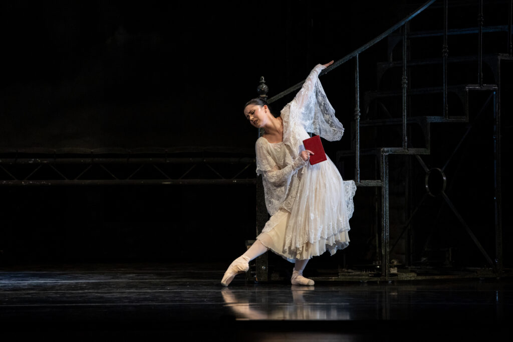 Alana Griffith, wearing a whit dress and long, lacy shawl, does a tendu with her right foot and pliés on her left as she hangs onto a bannister with her left hand and leans slightly towards her right. She holds a red book in her right hand.