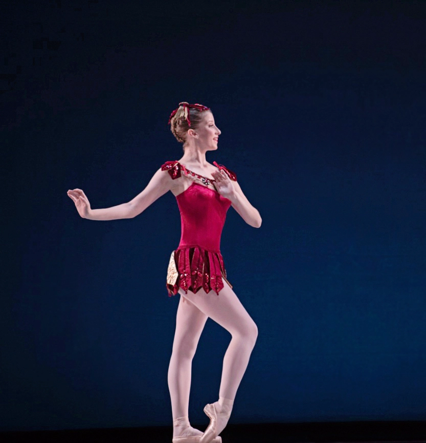 Prince wears a short red costume and stands in profile. Her feet are parallel and her front foot is pushed over her pointe shoe. She has one hand near her chest and the other extended gently to the side.