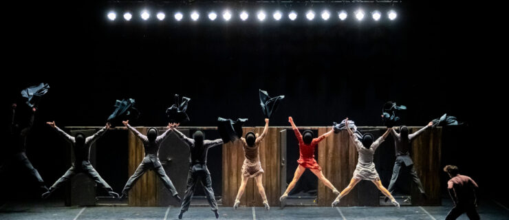 Eight dancers wearing black bowler hats face upstage and jump up, their legs and arms spread in a V shape. They all hold black sport coats in their right hands. A lone male dancer dances upstage left, facing upstage. A row of spotlights line the top of the stage's bacdrop.