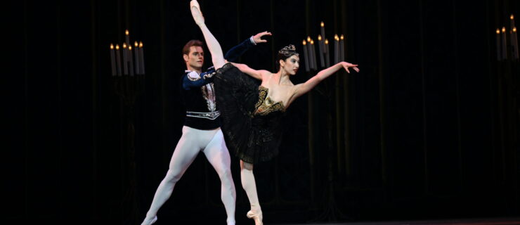 Chloe Misseldine and Michal Krčmář perform the Black Swan pas de deux onstage. Misseldine, wearing a black tutu with gold trim and a gold tiara, does a first arabesque penché on pointe with her right leg raised. Her partner, dressed in a black velvet tunic and white tights, stands behind her with his right leg in tendu side and holds her right hand.