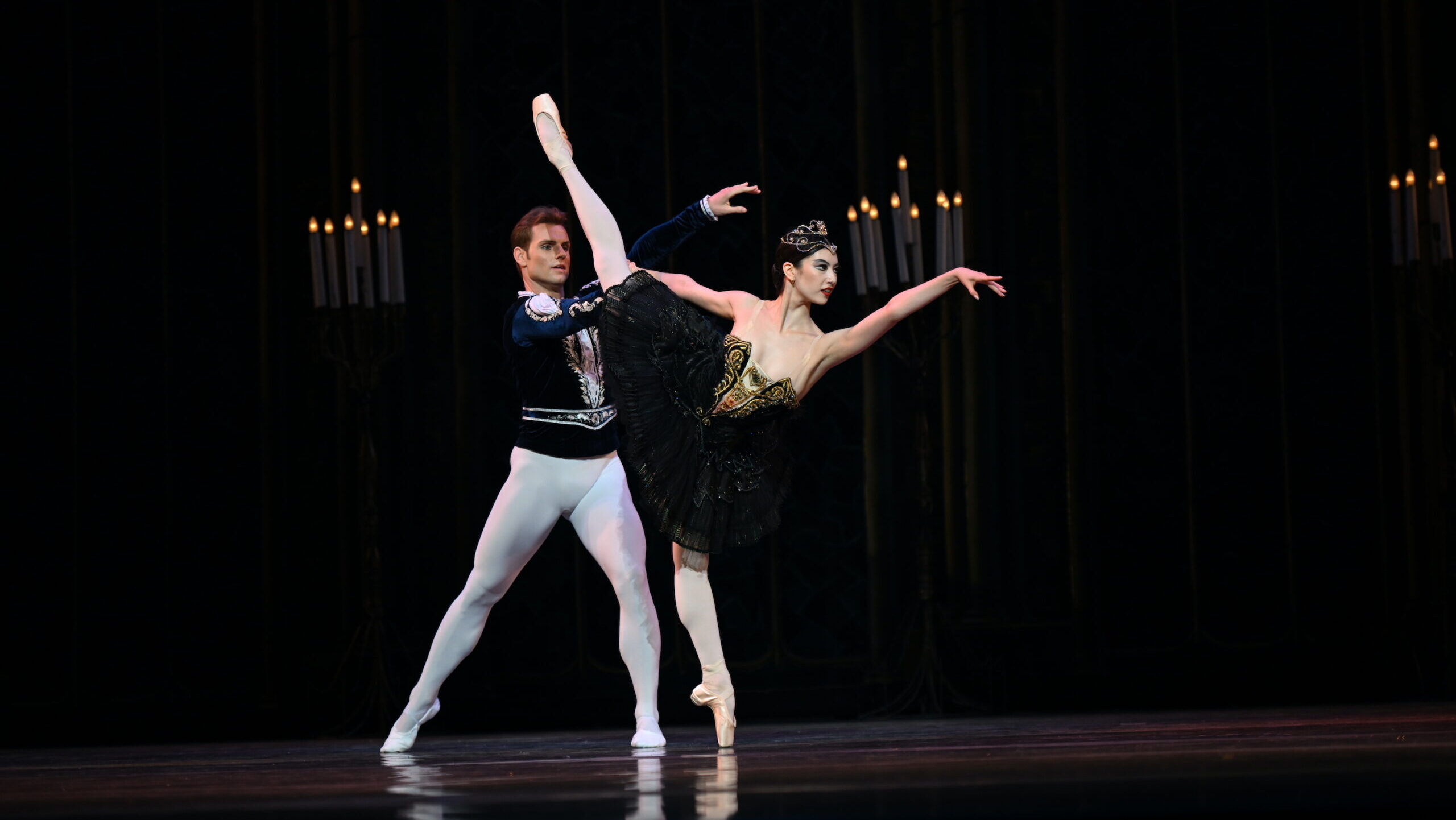 Chloe Misseldine and Michal Krčmář perform the Black Swan pas de deux onstage. Misseldine, wearing a black tutu with gold trim and a gold tiara, does a first arabesque penché on pointe with her right leg raised. Her partner, dressed in a black velvet tunic and white tights, stands behind her with his right leg in tendu side and holds her right hand.