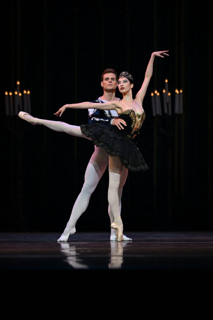 Chloe Misseldine and Michal Krčmář perform the Black Swan pas de deux onstage. Misseldine, wearing a black tutu with gold trim and a gold tiara, does a first arabesque on pointe with her right leg raised. Her partner, dressed in a black velvet tunic and white tights, stands behind her with hisright leg in tendu side and holds her by the waist.