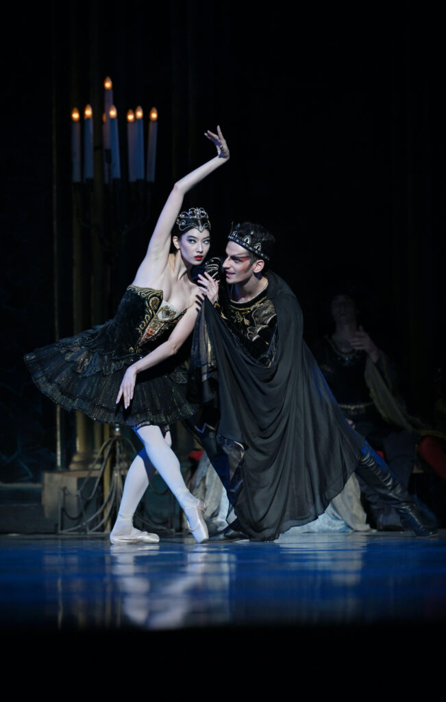 Chloe Misseldine stands on a bent left leg and pointe her right foot in tendu criosé as she lifts her right arm high next to her ear and crouches down to listen to the man next to her whisper in her ear. She wears a black tutu with gold trim and agold tiara, pink tights, and pointe shoes. The male dance next to her wears a large black cape, black and gold tunic, and large crown, as well as sinister makeup. They look like they are deviously plotting.