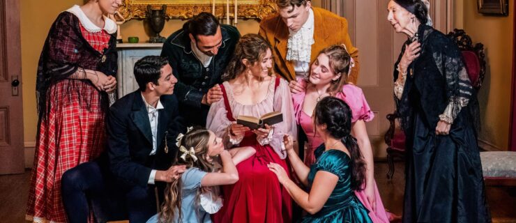 The cast of the Little Women Ballet gather around Jo March, who is seated and reading a book.