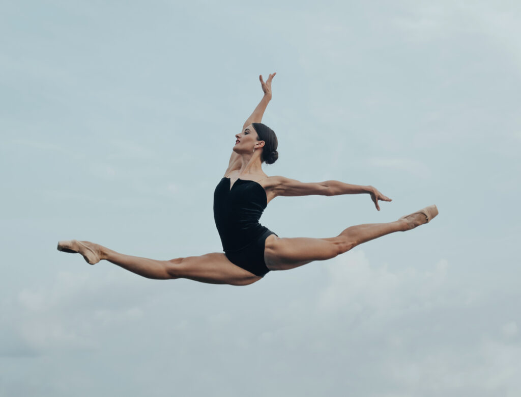 Evelina Godunova flies through the air in a saut de chat, suspended as if floating in front of a cloudy gray background. She wears a black leotard and pointe shoes, her dark hair slicked into a low bun.