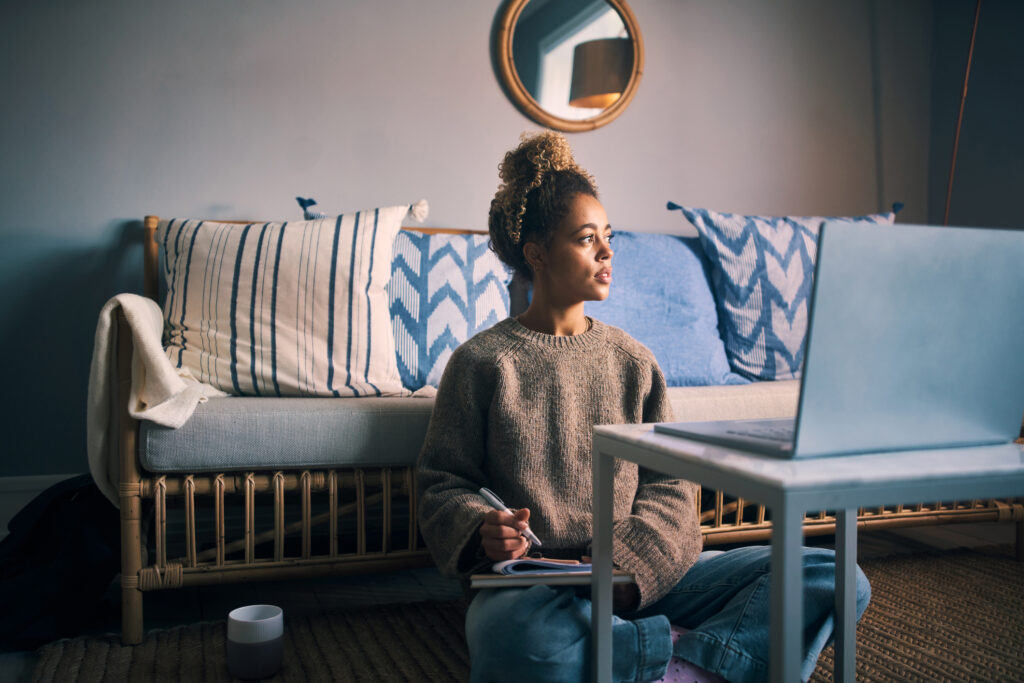 A young woman looks away thoughtfully while writing notes and using a laptop at home.