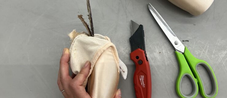 A woman's hand is shown holding a pointe shoe, pulling back the back of it to expose the shank. A red box cutter and pair of scissors lie to the right of her hand on a gray dance studio floor.