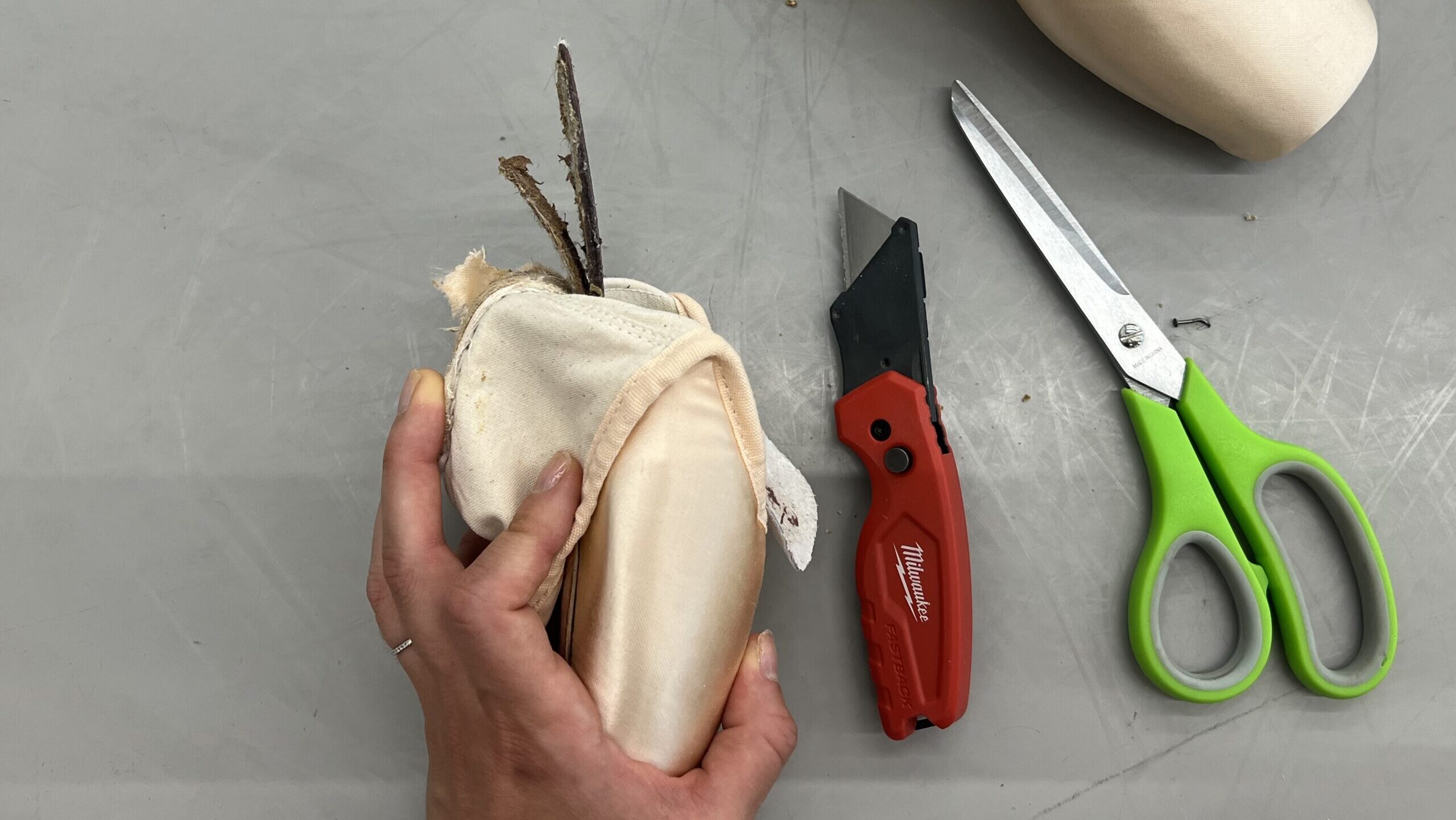 A woman's hand is shown holding a pointe shoe, pulling back the back of it to expose the shank. A red box cutter and pair of scissors lie to the right of her hand on a gray dance studio floor.