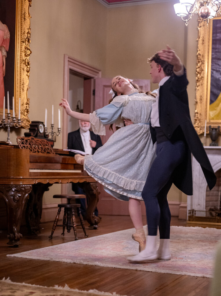 A dancer portraying Amy March, wearing a blue dress and pointe shoes, stands en pointe with her back leg in attitude. She is held by a dancer playing Laurie. They stand on an antique carpet in a Victorian-era room.