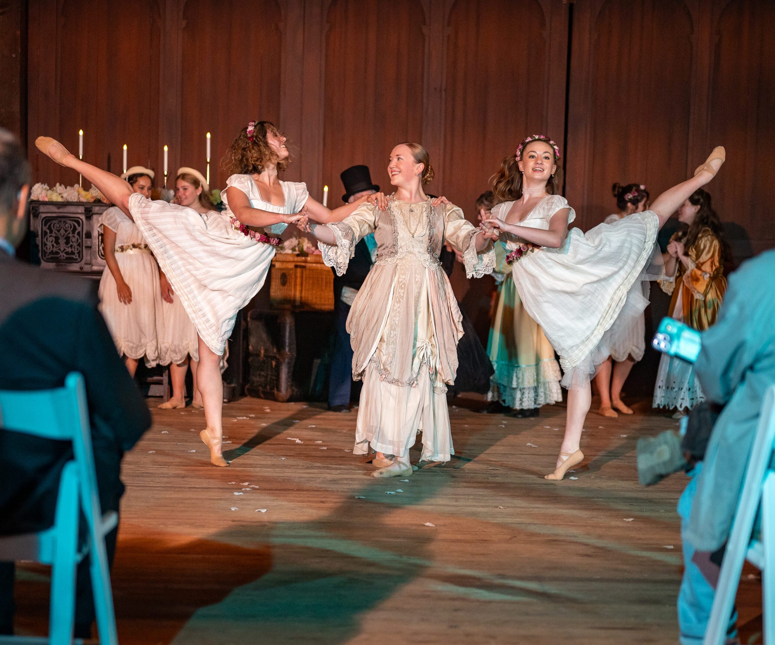 A woman in a Victorian dress supports a dancer in arabesque on each arm.