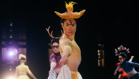 Jacob Seltzer wears an elaborate yellow and white costume reminiscent of a Frida Kahlo painting. He faces profile, his head turned toward the camera as he lifts his long skirt and dances onstage.