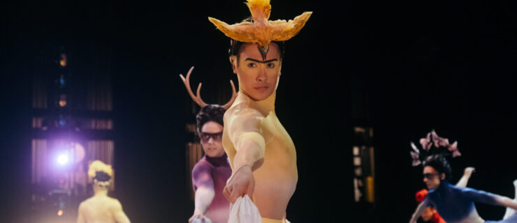 Jacob Seltzer wears an elaborate yellow and white costume reminiscent of a Frida Kahlo painting. He faces profile, his head turned toward the camera as he lifts his long skirt and dances onstage.