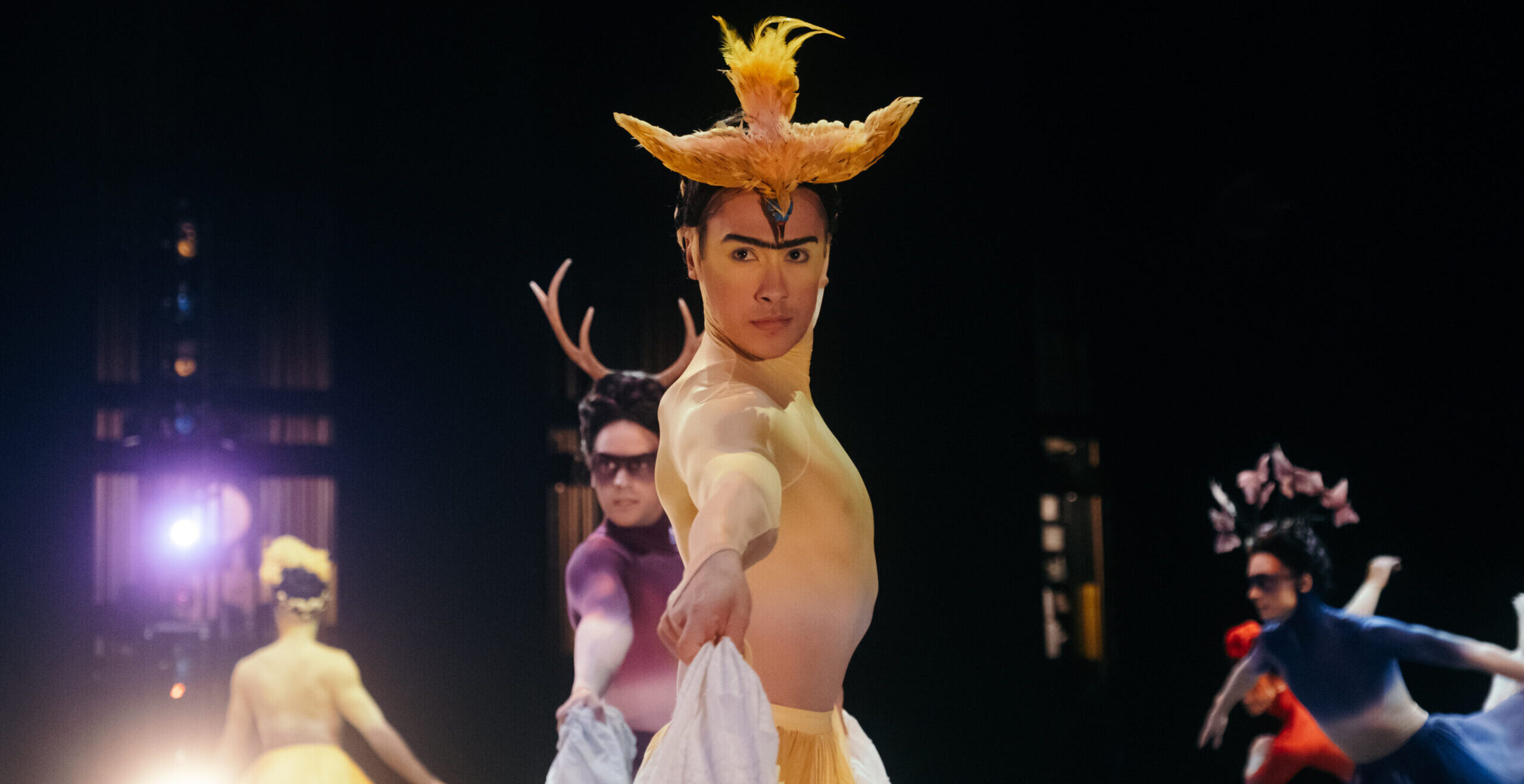 Jacob Seltzer wears an elaborate yellow and white costume reminiscent of a Frida Kahlo painting. He faces profile, his head turned toward the camera as he lifts his long skirt and dances onstage.