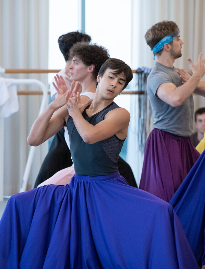 Jacob Seltzer wears a long blue skirt in rehearsal, his hands poised to clap as he does a deep grand plié in second. He is surrounded by other dancers in long blue, purple, and pink skirts.