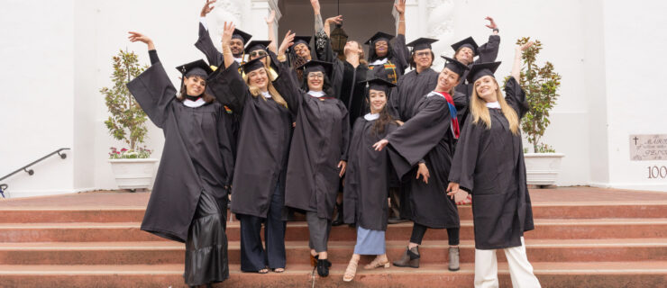 A group of dancers in graduation caps and gowns cluster together on a small staircase, posing and smiling triumphantly.