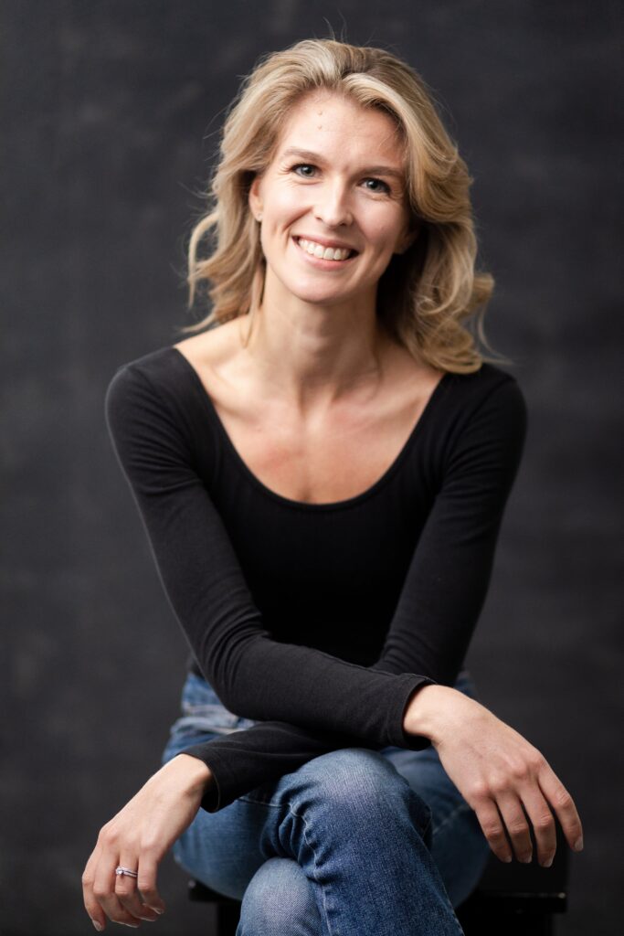 Lucy Ashe sits down on a stool, facing directly front, and crosses her right leg over her left. She crosses her forearms and rests them on her thighs. She has shoulder length blonde hair and wears a form-fitting, long-sleeved black shirt and blue jeans, and looks drectly at the camera with a large smile.