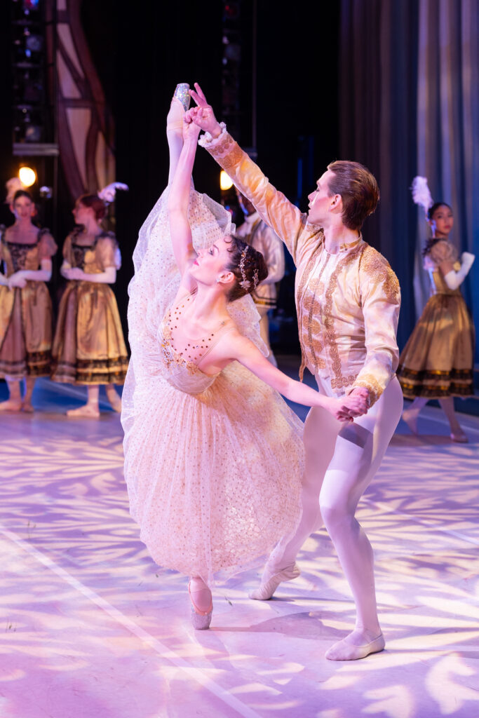 Marie Harrison-Collins, dressed as Cinderella in a light gold tulle ballgown, does a penche on pointe as her partner,  Josiah Cook supports her by holding her hands. He wears a gold and white prince's costume and lunges with Marie, his right leg in tendu a la seconde. Textured purple light fills the stage, and a corps of ball guests surround them in the background.