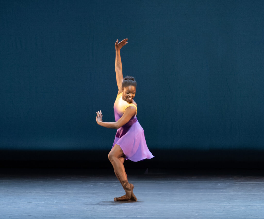 Michaela Mabinty DePrince stands onstage on her right foot, her body facing stage right, as she points her left foot in parallel and sweeps her arms up to her right. She looks over her left should and down at her feet with a large smile. She wears a purple, knee-length dress and brown pointe shoes.