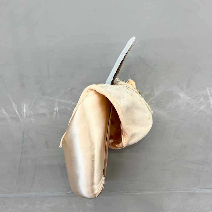 A new pointe shoe is shown on its side, lying on a dance studio floor. The outsole is pulled back, exposing the insole and shank.