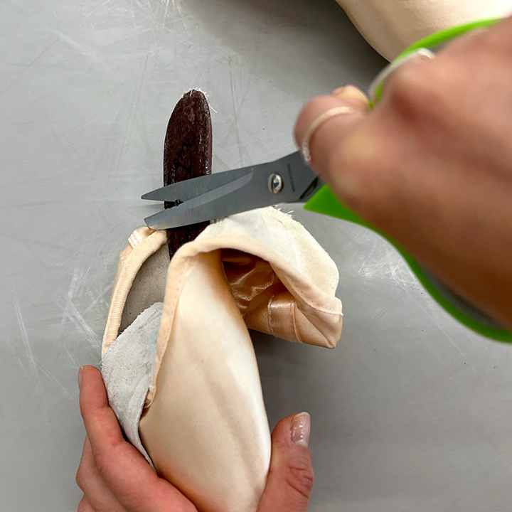 A woman's hands are shown close up as she holds a pointe shoe with her left hand, with the sole pulled back to expose the shank. With her right hand, she grasps a nail in the shank with a pair of scissors.