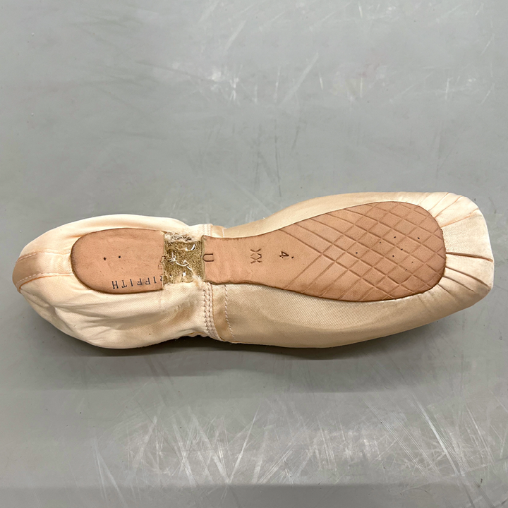 A pointe shoe is shown lying fce down on a gray dance studio floor. A rectangular chunk of the sole has been cut out where the heel meets the arch.