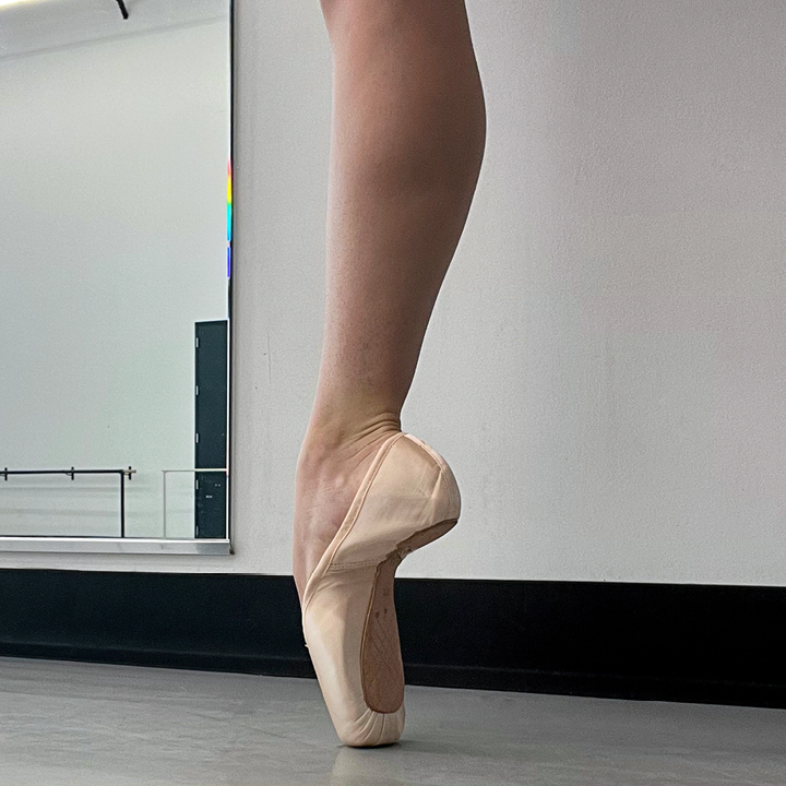 A ballerina's leg is shown from the knee down, standing on pointe in a new pointe shoe.