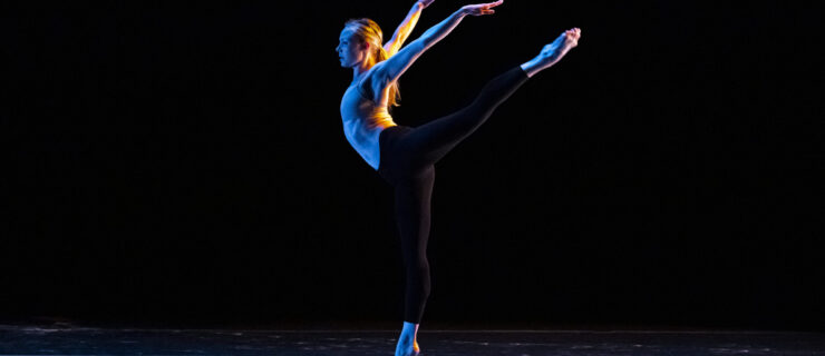 Sara Mearns wears a nude leotard and black tights. In dark lighting onstage, she does an arabesque facing profile, her arms lifted in a wide V behind her.