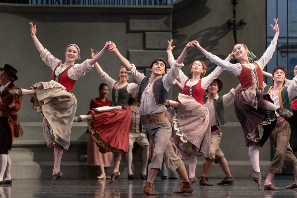 A large corps de ballet performs onstage in peasant costumes, holding hands in three long lines across the stage. Jacob Seltzer lunges in the center with a proud expressoin.