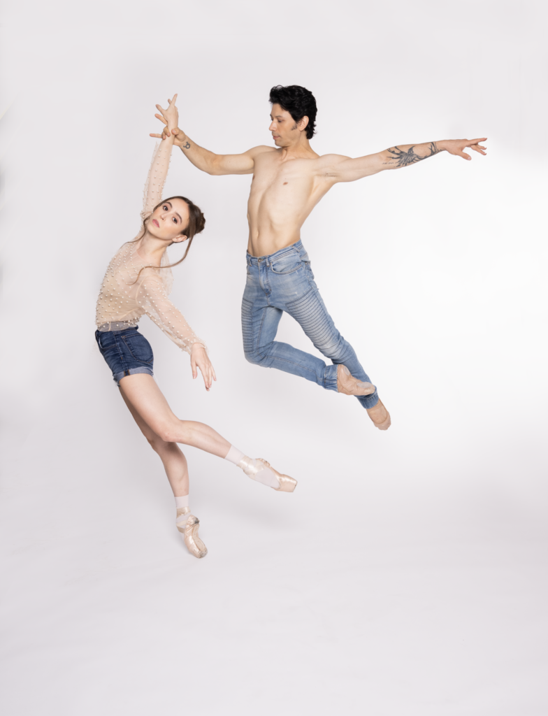 Skylar Brandt and Herman Cornejo dance together in front of a light gray background. Brandt wears jean shorts, a sheer nude longsleeve top with pearls, white socks, and pointe shoes. Cornejo wears jeans and ballet shoes. Brandt leans forward in a low attitude derriere on pointe, her right arm up and left extended to the side. Cornejo holds her right wrist as he jumps in cou de pied devant, extending his left arm out to the side.