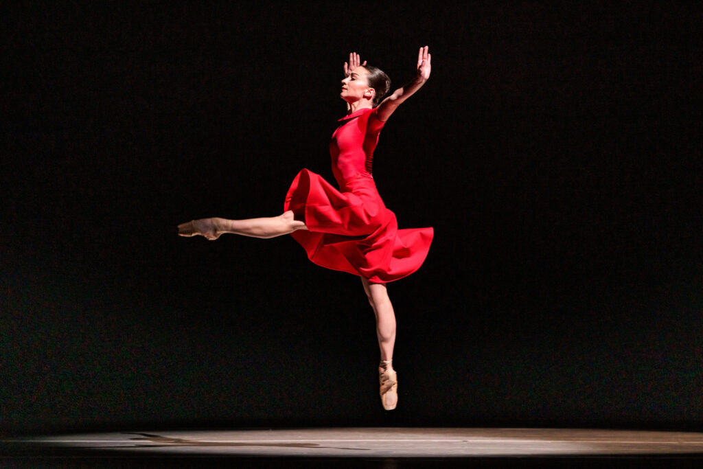 Terez Dean Orr is shown jumping in profile during a performance. She wears a knee-length red dress and is spotlit from above. Facing stage right, she jumps off of her right foot and kicks her left leg at 90 degrees in front of her, with her arms straight out to her sides and her wrists flexed.