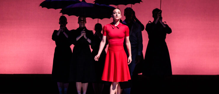 Terez Dean stands in fifth position center stage, her arms at her sides as she faces directly front and gazes out into the audience. She wears a knee-length, short-sleeved red dress and pink pointe shoes. Behind her, in the shadows, five dancers stand in parallel, holding parasol umbrellas.