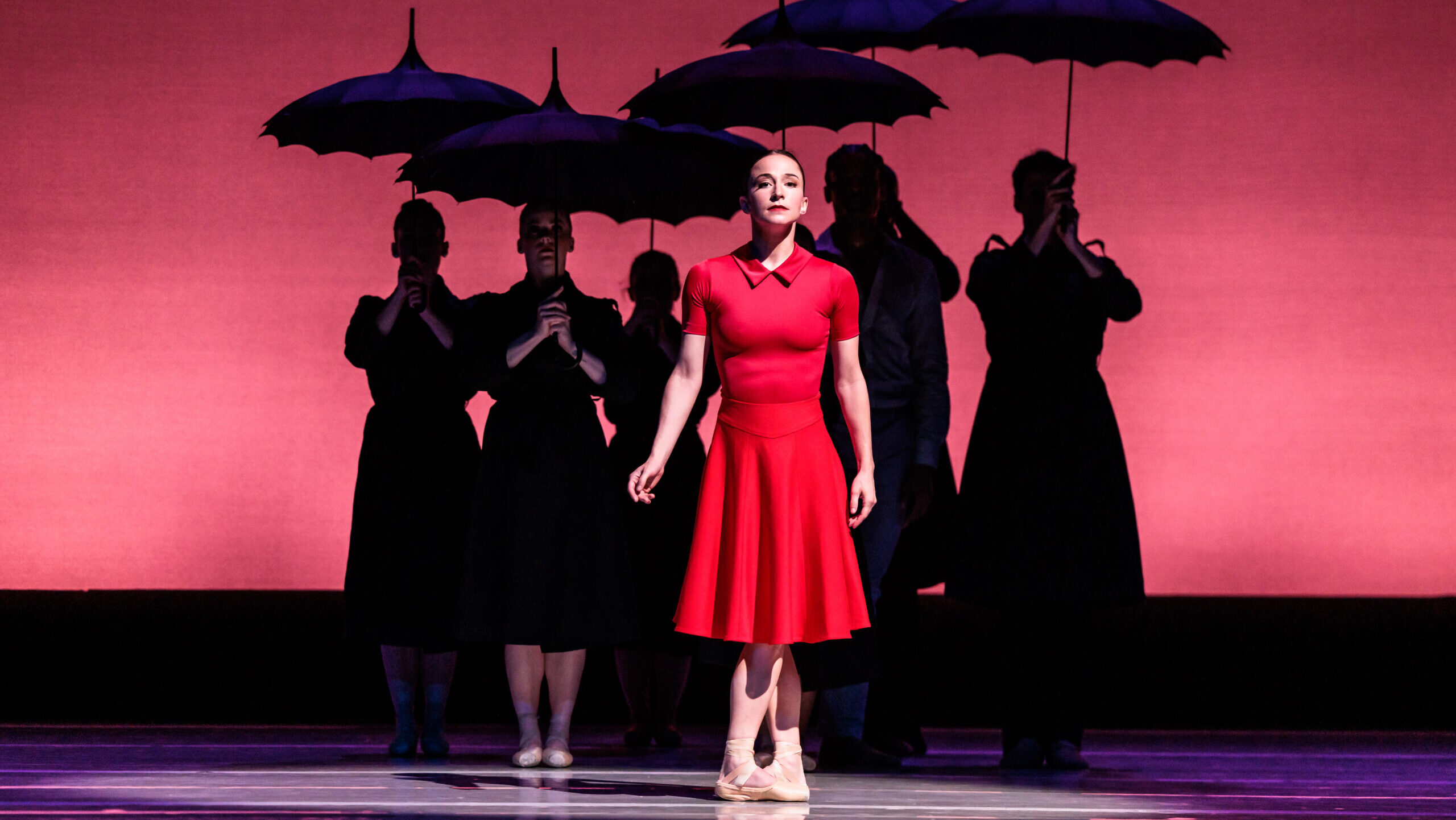 Terez Dean stands in fifth position center stage, her arms at her sides as she faces directly front and gazes out into the audience. She wears a knee-length, short-sleeved red dress and pink pointe shoes. Behind her, in the shadows, five dancers stand in parallel, holding parasol umbrellas.