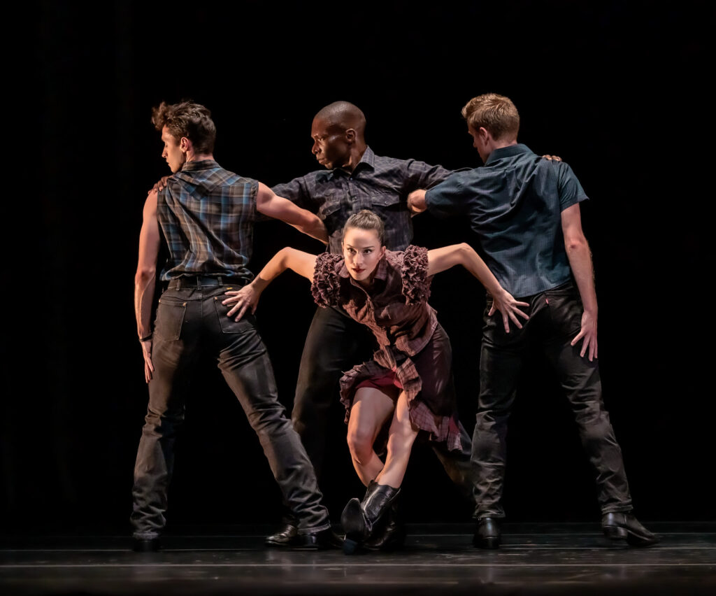 Terez Dean Orr, in black cowboy boots and a borwn, frilly dress, sits back on her right heel onstage and places her hands on the rear ends of two male dancers on either side of her. They face the back of the stage and look to their left, while another man in between them and behind Dean faces front and turn his head to his right. The men wear plaid button down shirts, black jeans, and black boots.