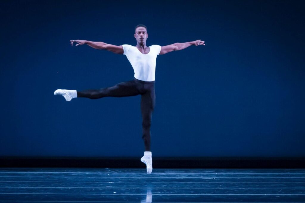 Joshua Bodden suspends mid-air in a sauté a la seconde, his right leg extended 90 degrees and his arms extended in a straight line with his shoulders, parallel to the stage floor. He wears a fitted white shirt, black tights, and white socks with white ballet shoes. The backdrop is a deep blue.