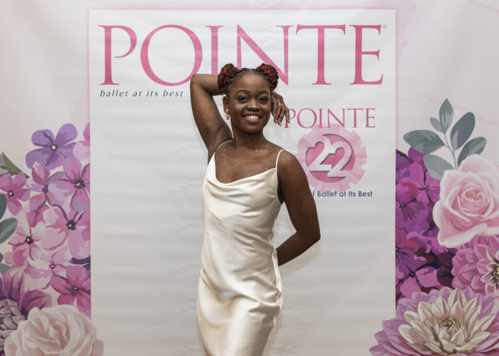 Michaela Mabinty DePrince wears a silky white gown and poses in front of a step-and-repeat with the Pointe magazine logo across the top. Her right arm is bent and placed behind her head while her left arm is down and behind her body. She smiles brightly.