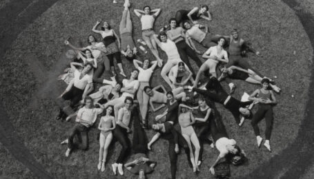 In this black and white image, a large company of ballet dancers in rehearsal clothing form a circular shape and lay on the grass casually and look up towards the camera as it photographs them from above.