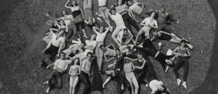 In this black and white image, a large company of ballet dancers in rehearsal clothing form a circular shape and lay on the grass casually and look up towards the camera as it photographs them from above.