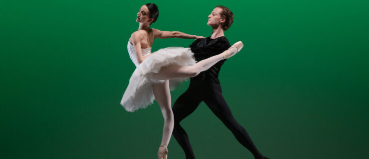 In front of an emerald green backdrop, a female dancer in a white classical tutu stands in attitude derriere on pointe, supported by her partner, who lunges behind her and holds her waist. He wears a black unitard.