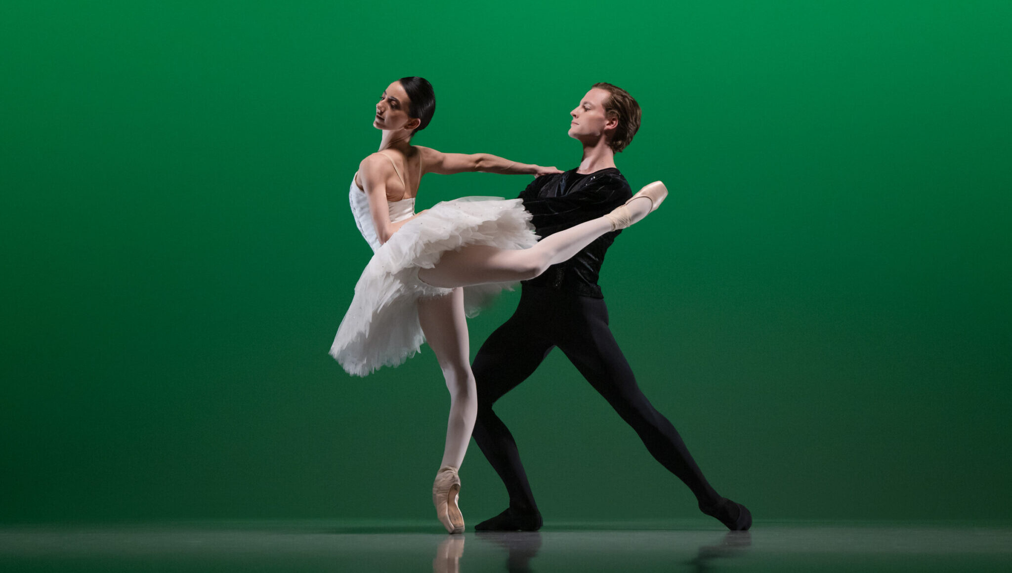 In front of an emerald green backdrop, a female dancer in a white classical tutu stands in attitude derriere on pointe, supported by her partner, who lunges behind her and holds her waist. He wears a black unitard.