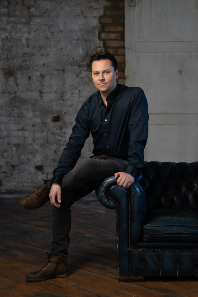 Christopher Marney, wearing a dark button down shirt and gray jeans, sits on the arm of a black leather chair in front of a stone and brick background. He crosses his legs and looks into the camera pensively.