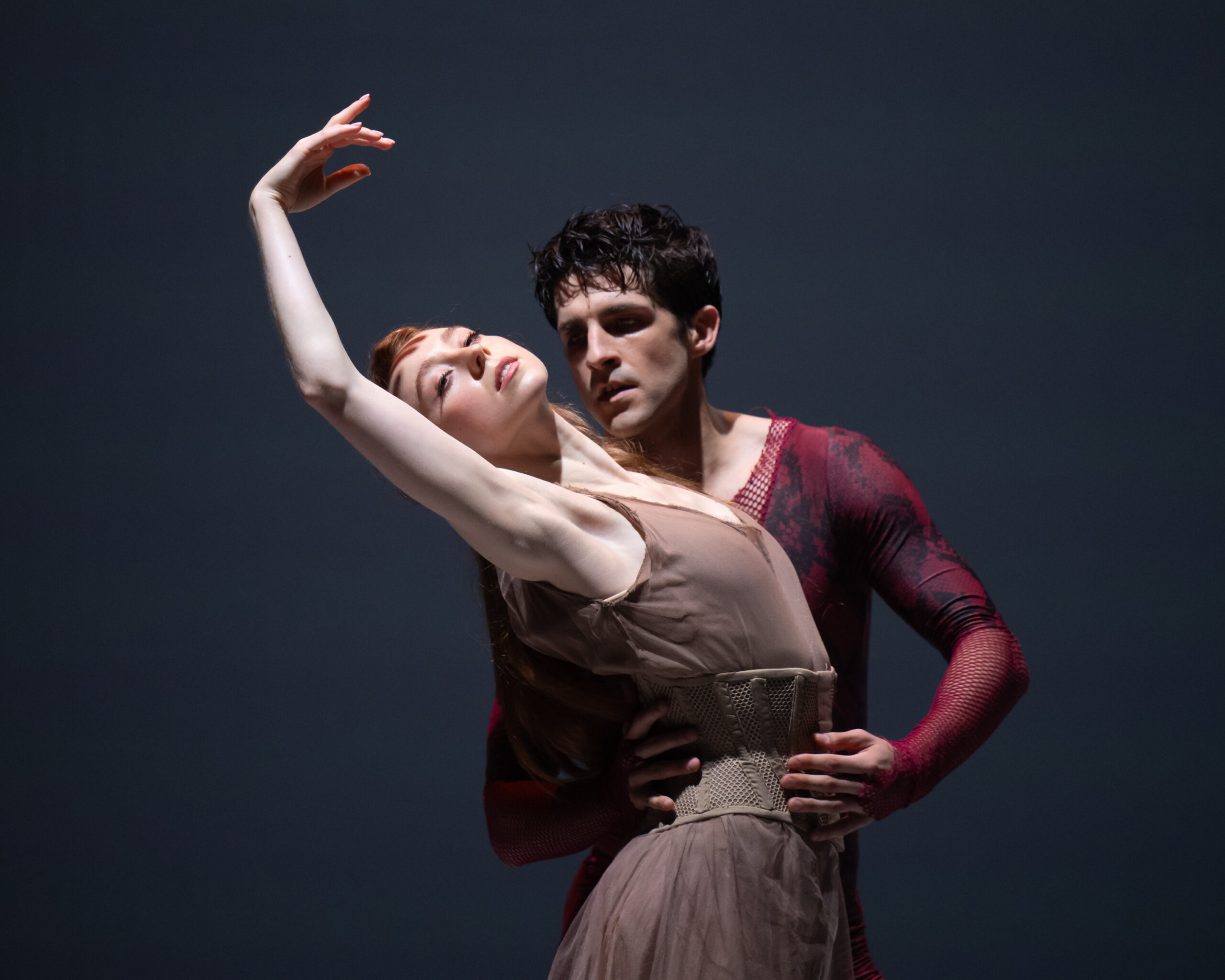 A female dancer in a brown-gray dress arches back with her right arm in high fifth. Behind her, her parter holds her waist and looks at her intently. He wears a red and gray textured unitard.