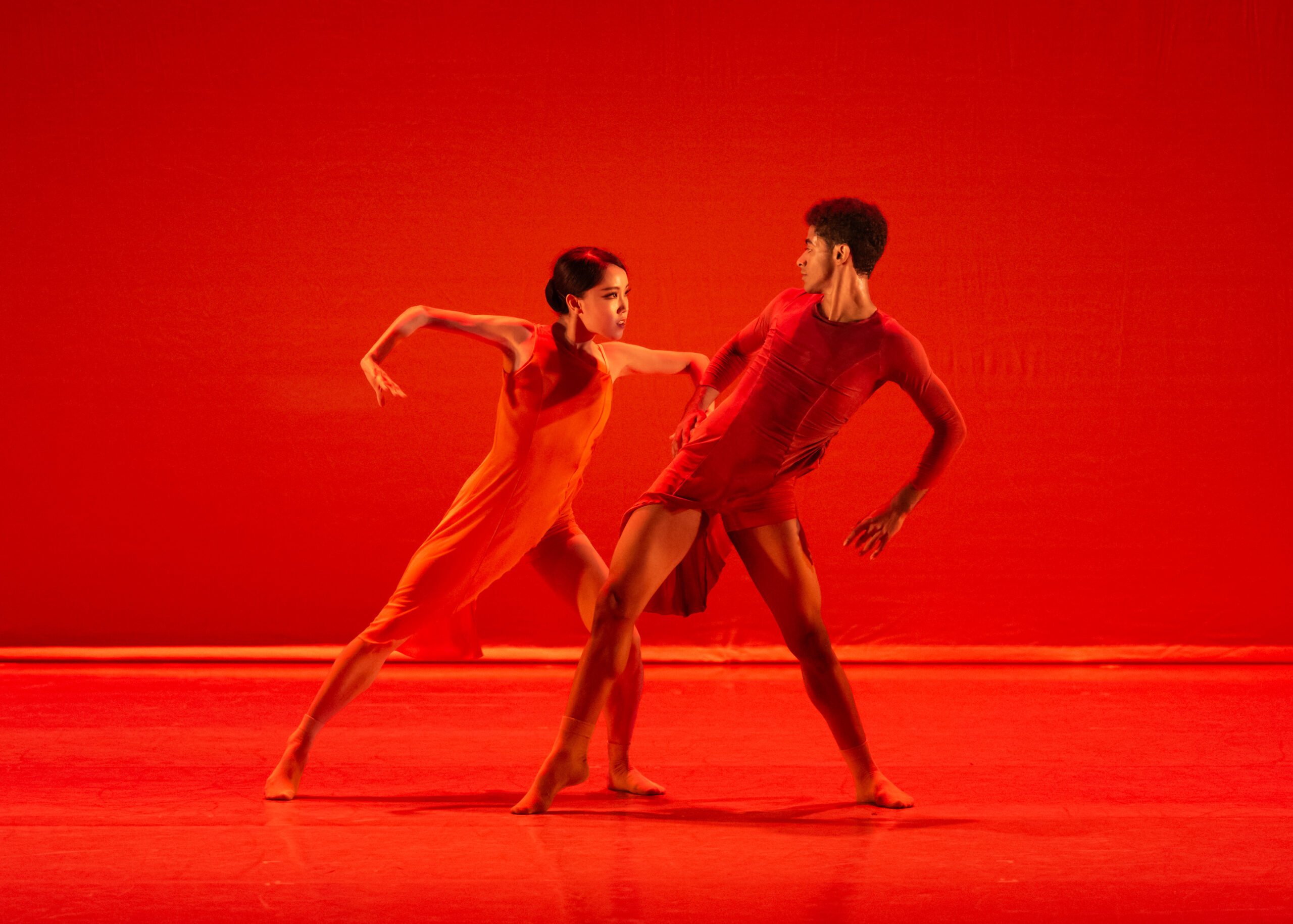 Two dancers in red costumes dance together onstage, lit by deep red lighting. On the left, a female dancer lunges toward the male dancer on the right, her arms extended and bent in a strong, angular position. The male dancer leans back, his arms bent behind him.