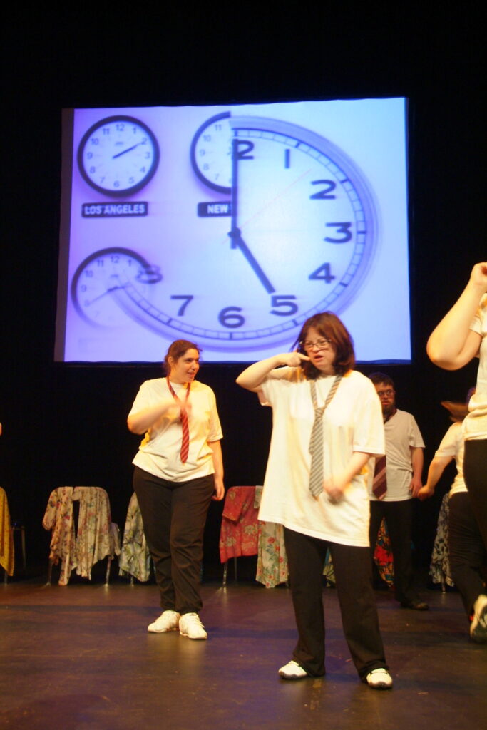 Louise Forster, wearing an oversized white t-shirt, black yoga pants, sneakres and loose-fitting tie, stands onstage during a performance and points to the left with her right hand, looking at her finger intently. Other dancers in similar costumes move behind her. A large projection showing a clock is shown behind them onstage.