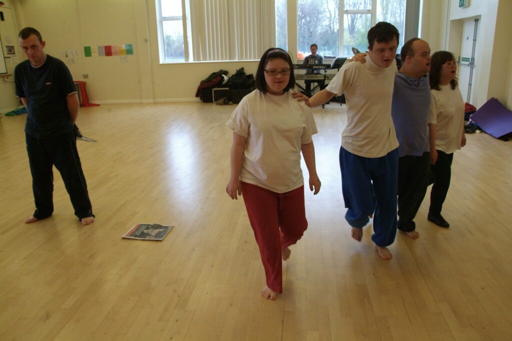 Louise Forster, wearing a white t-shirt and red pants, walks forward in a sunny dance studio. To her left, a line of three other dacers, linked shoulder-to-shoulder walk slightly behind her while the one closest to her places his hand on her shoulder.