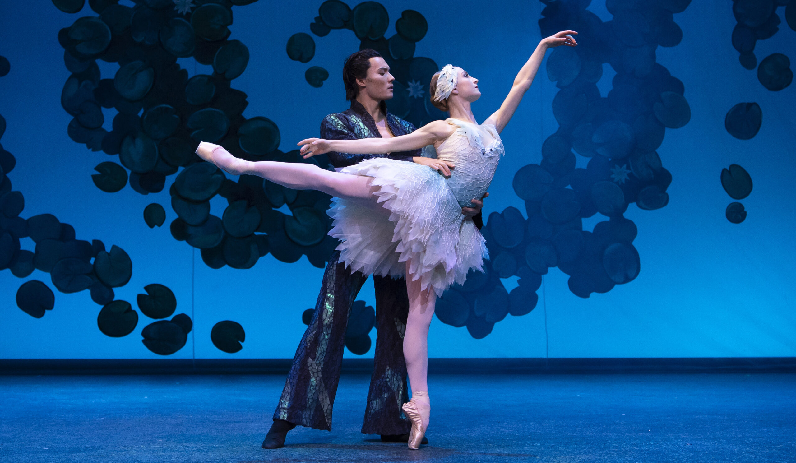 In front of a blue background with a dark textured swirl, a male dancer in a long dark, shimmering suit partners a dancer dressed as Odette, in a tattered white dress and crown. She stands in arabesque on pointe.